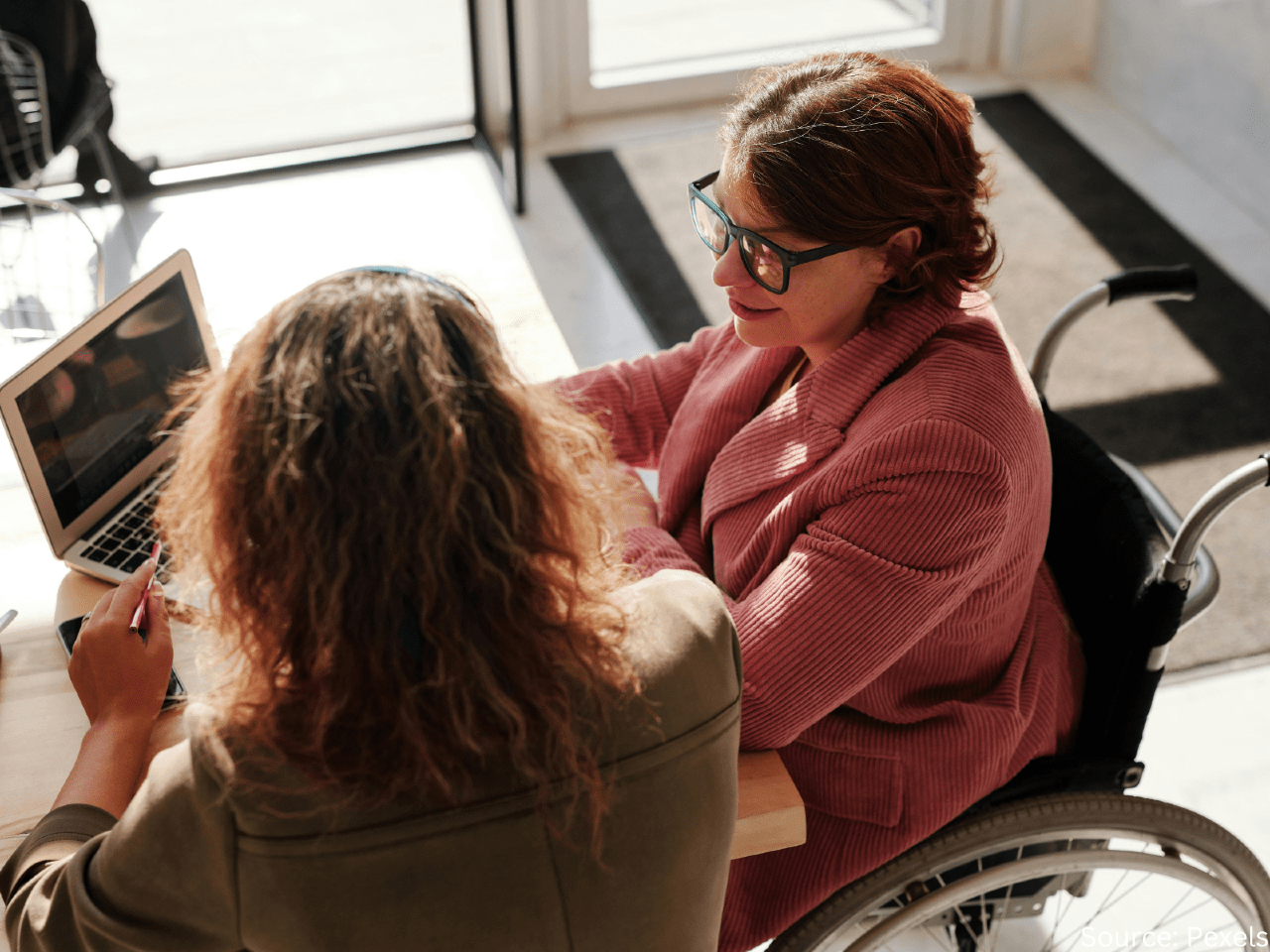 a woman in a wheelchair speaks with another woman using a laptop